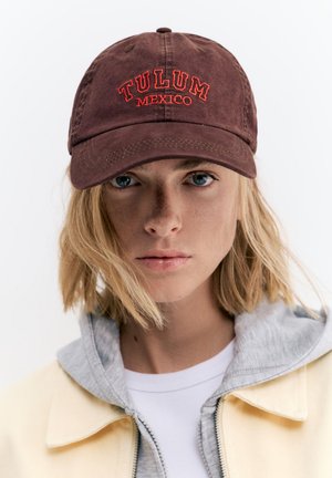 Ung person med blondt hår iført en brun kasket med teksten "TULUM MEXICO", en grå hættetrøje og en lys gul jakke mod en ensfarvet baggrund.