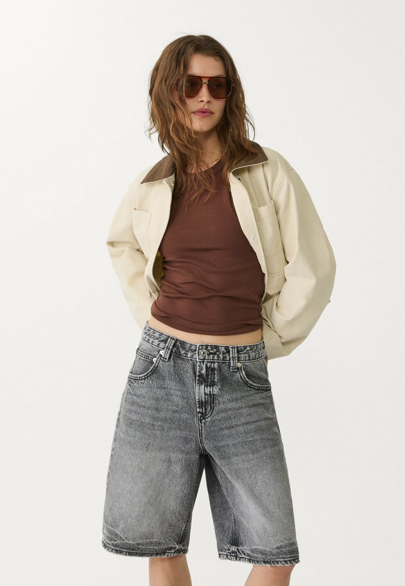 Veste crème avec col marron, débardeur marron ajusté et shorts en denim noir délavé de mi-longueur. Le modèle porte des lunettes de soleil marron.