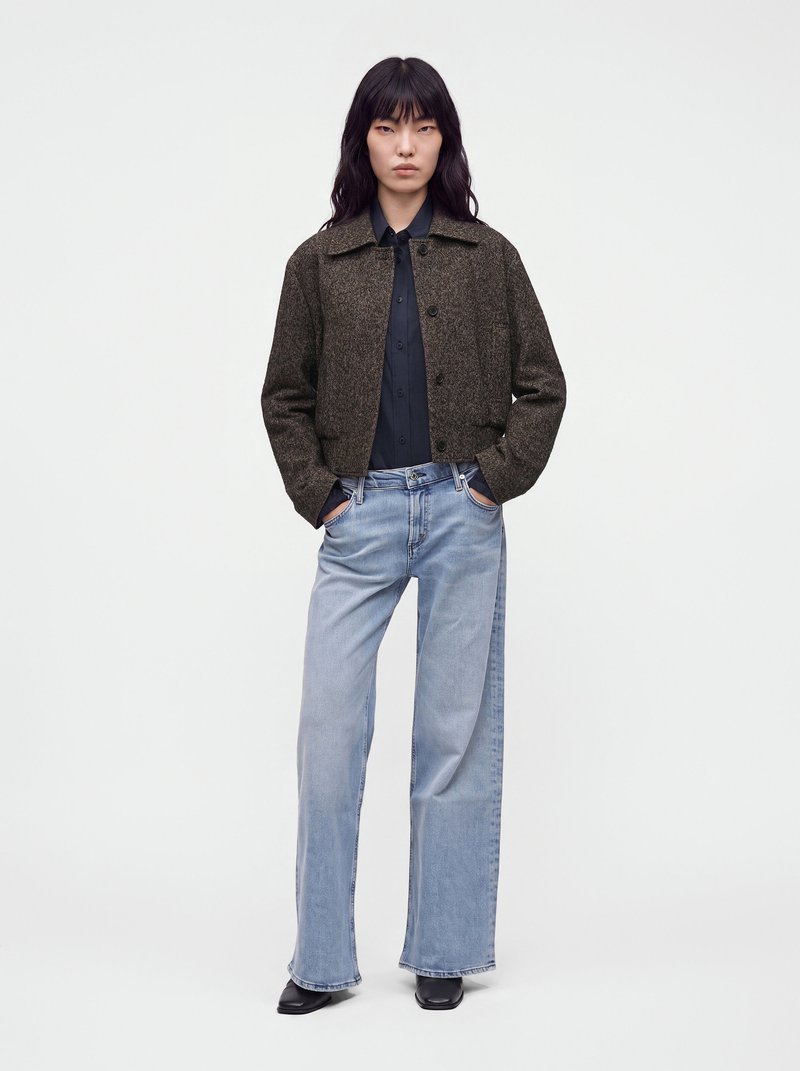 Femme portant une veste marron foncé boutonnée, une chemise noire, un jean large bleu clair et des chaussures noires, debout les mains dans les poches.
