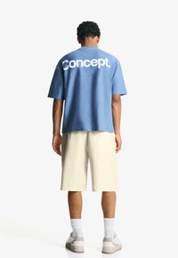 T-shirt blu a maniche corte con logo "Concept." in bianco sulla schiena, abbinato a pantaloni corti beige. Vestibilità casual; il tessuto appare morbido.