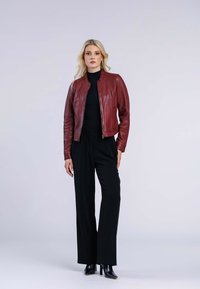 Veste en cuir rouge avec fermeture éclair, col rond et manches longues. Associée à un pantalon large noir et des bottes à talons. Fond uni.