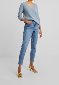 Maglione azzurro chiaro a maglia con scollo a V, abbinato a jeans in denim blu a vita alta e sandali gialli con cinturini. Outfit semplice e casual.