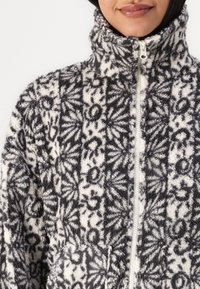 Veste en polaire noire et blanche, avec un col haut, fermeture éclair et texture à motifs floraux. Comprend une poche avant.