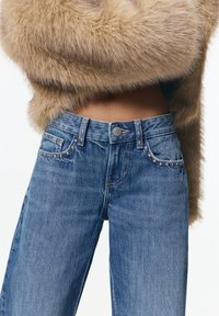 Jean en denim bleu avec une taille mi-haute, des coutures détaillées et des clous décoratifs sur les poches. Le tissu a une texture douce.