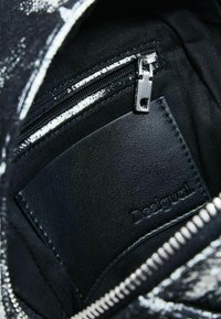 Interiorul din țesătură neagră al unei genți, având un buzunar cu fermoar și un eticheta din piele neagră netedă, cu sigla „Desigual” embosată. Hardware-ul fermoarului este de culoare argintie.