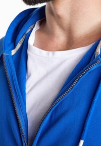 Sweat à capuche zippé bleu avec un t-shirt blanc en dessous. Le sweat présente une texture douce, une fermeture éclair visible et des détails en cordon.
