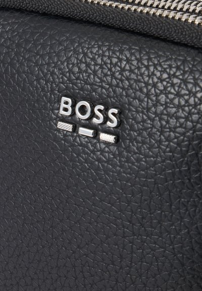 Gros plan sur un cuir noir texturé avec le logo BOSS argenté et une partie d'une fermeture éclair argentée en haut.
