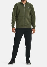 Sudadera con cremallera color verde oliva con dos bolsillos delanteros, puños acanalados y pantalones deportivos negros. Logotipo blanco en el pecho y en el lateral de los pantalones.