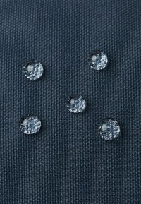 Transparente Wassertropfen auf dunkel strukturiertem Stoff, fünf Tropfen variieren leicht in der Größe, mit klaren Reflexionen und ohne sichtbare Verunreinigungen.