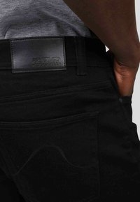 Pantalons en denim noir avec un patch en cuir à l'arrière présentant des éléphants en relief. Le tissu est lisse, avec des détails de couture subtils.