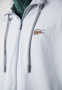 Chaqueta gris claro con cierre de cremallera, un logo de cocodrilo bordado y detalles con cordones, fabricada con un tejido suave y resistente al agua.