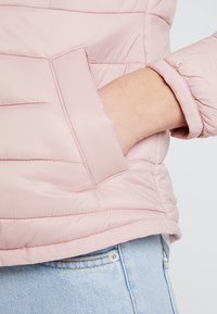 Lys pink puffjakke med en quiltet struktur, der har en sidelomme og en blød, fleecefôret manchet, parret med lyseblå denim.