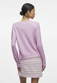 Jasnoróżowy sweter z dzianiny z prążkowanym dołem i mankietami, noszony na tle teksturowanej jasnoróżowej spódnicy z poziomymi falbankami.