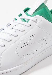 Baskets blanches avec doublure intérieure verte, lacets blancs, logo de marque perforé sur le côté, et languette en tissu verte à l'avant.