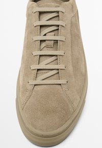 Beige suede sneaker with flat laces, textured upper, rounded toe, and a rubber sole. Minimalist design with stitched detailing.