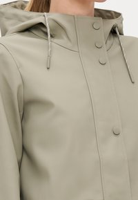 Chaqueta impermeable beige con capucha, que cuenta con botones a presión, un cierre frontal con cremallera y cuerdas de ajuste en contraste para un ajuste personalizado. Textura suave.