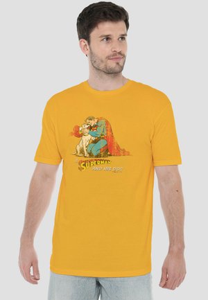 Mann trägt gelbes T-Shirt mit Vintage-Grafik von Superman, der kniet und einen Hund umarmt, Beschriftung lautet „Superman und sein Hund.“