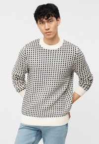 Jeune homme aux cheveux courts et bouclés portant un pull blanc à motifs bleus marins et un jean bleu clair, debout, les mains derrière le dos.