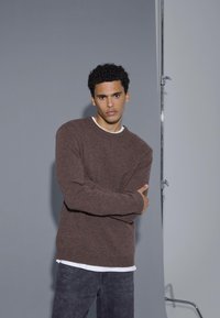 Pull en maille marron avec un col rond, porté sur une chemise blanche, associé à un jean foncé. Le fond neutre met en valeur les détails de la tenue.