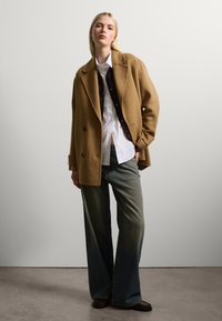Cappotto doppiopetto in misto cammello sopra una camicia bianca e jeans larghi in denim. Texture liscia con tasche laterali e decorazioni con bottoni.