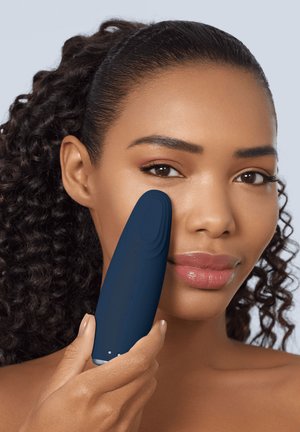 Femme aux cheveux bouclés tenant un appareil de nettoyage facial bleu foncé contre sa joue droite, démontrant l'utilisation pour les soins de la peau.