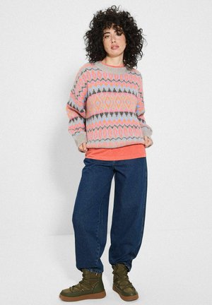 Sweter w jasnym różu z geometrycznymi wzorami w odcieniach szarości, niebieskiego i pomarańczowego; luźny krój. Połączony z ciemnoniebieskimi spodniami o szerokich nogawkach i oliwkowymi botkami.
