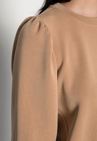 Top beige de manga larga con textura acanalada y mangas abullonadas con fruncido. La tela es suave y con un ligero brillo. Diseño de escote sencillo.