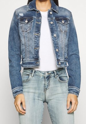 Femme portant une veste en jean bleu délavé sur une chemise blanche, associée à un jean bleu clair, debout devant un fond uni.