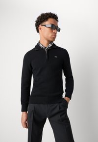 Mann trägt schwarzen Zip-up-Pullover, Hemd mit floralem Kragenmuster, schwarze Hose und rechteckige dunkle Sonnenbrille, steht vor weißem Hintergrund.
