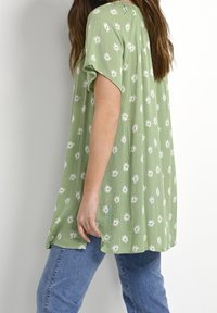 Zielony top z krótkim rękawem o pogniecionej fakturze i białym kwiatowym wzorze, zestawiony z niebieskimi dżinsami. Dopasowany okrągły dekolt i luźny krój.