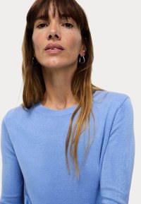 Ljusblå stickad tröja med rund halsringning, långa ärmar och en mjuk textur. Håret är långt och rakt, med en naturlig look.