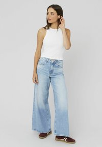 Jeune femme debout, portant un haut sans manches blanc, un jean large bleu clair et des baskets bordeaux avec des rayures blanches.