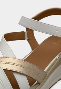 Λευκό σανδάλι με ρυθμιζόμενους δερμάτινους ιμάντες και χρυσή μεταλλική λεπτομέρεια. Η σόλα έχει μαλακή επένδυση με λείο καφέ εσωτερικό.