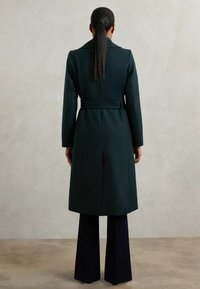 Pikendusega naine, kellel on pikk hobusesaba, kannab tumerohelist vööga mantlit ja musti laia jala pükse, seistes ühetaolisel heledal taustal.