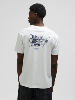 Hombre con camiseta blanca con ilustraciones en azul marino de un jugador de tenis y texto "Esto me dio una lección" en la espalda.