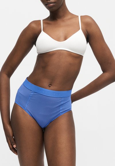 Γυναίκα που φοράει λευκό σουτιέν και μπλε ριγέ εσώρουχο με ψηλή μέση και ζώνη με λογότυπο, ποζάρει σε απλό φόντο.