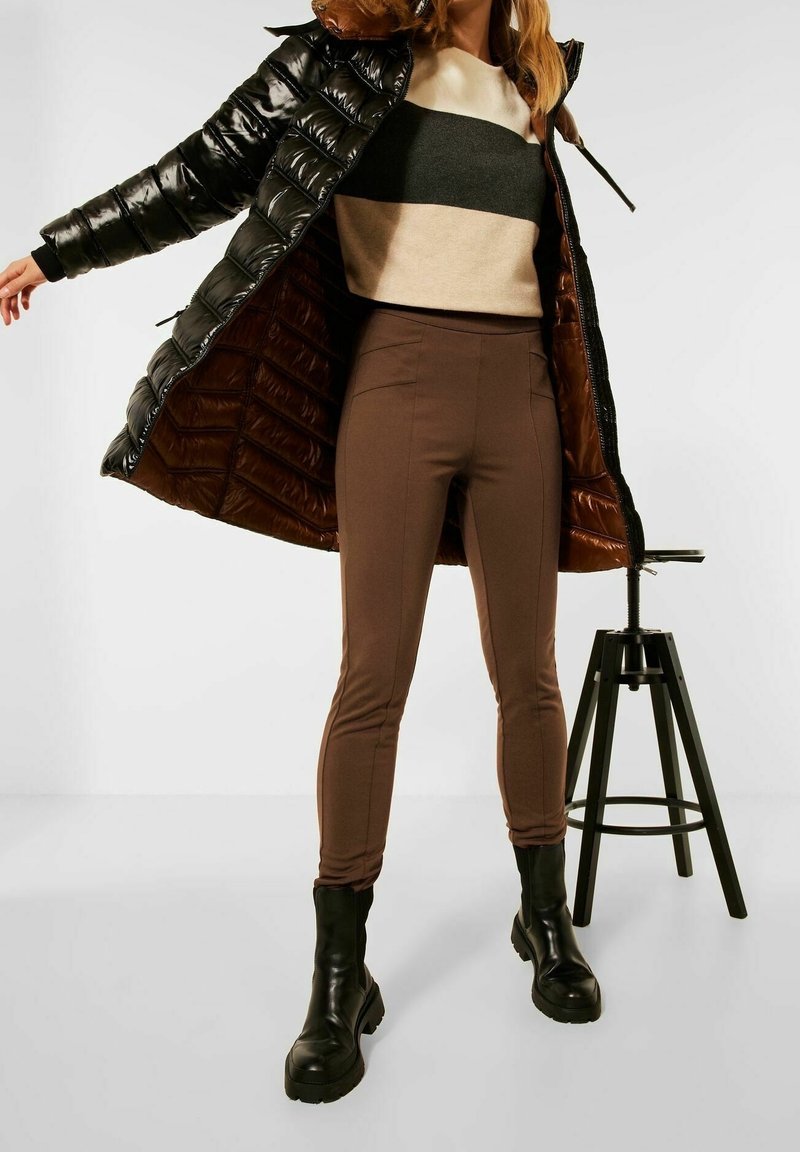 Kvinna klädd i en svart glänsande pufferjacka med brun interiör, bruna högmidjade byxor, svarta stövlar och en beige- och svartrandig tröja.