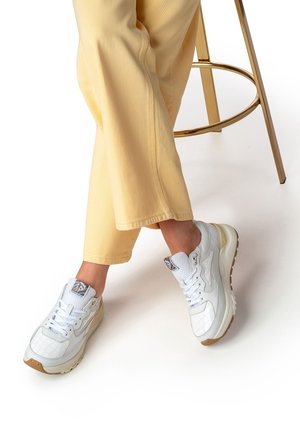 Zapatillas blancas con textura y acabado brillante, suela de goma y cordones, combinadas con pantalones vaqueros amarillos, sobre un fondo blanco.