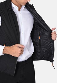 Gilet nero con chiusura a zip e accenti arancioni a contrasto. L'interno presenta un motivo trapuntato e una tasca aggiuntiva con zip.