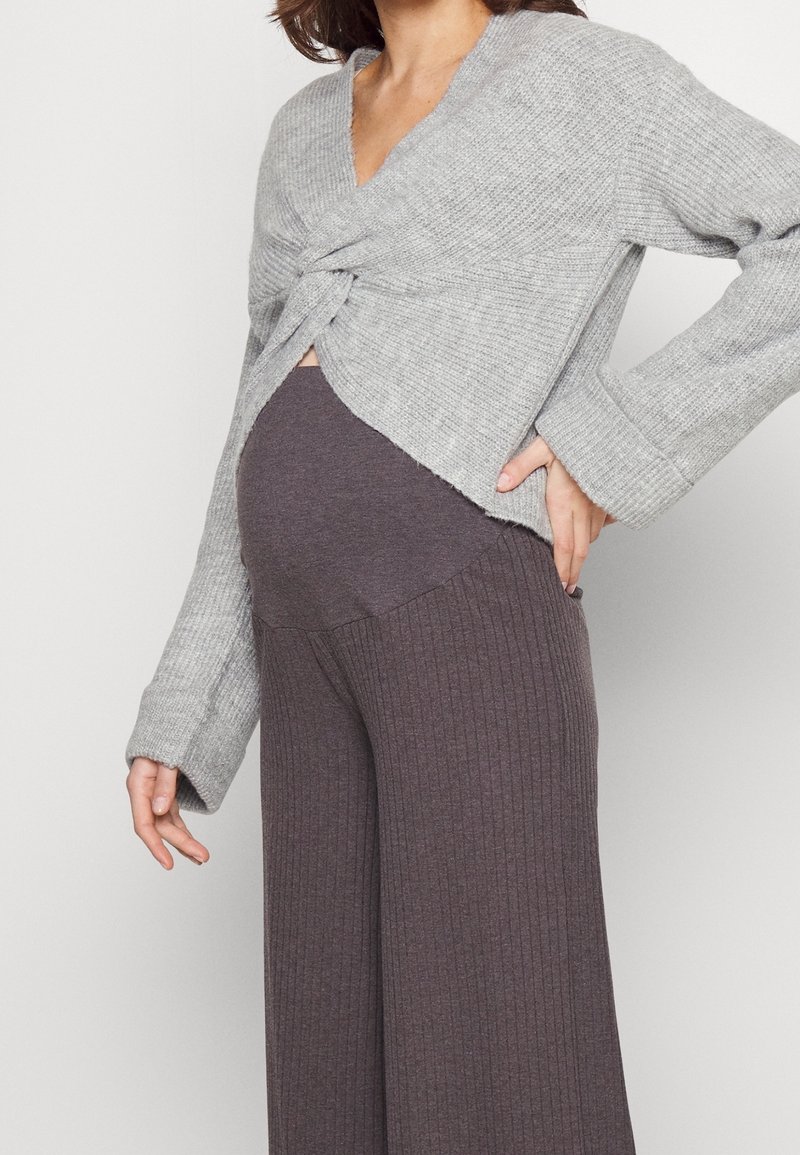 Femme enceinte portant un pull gris torsadé et un pantalon de maternité côtelé gris foncé, main dans le bas du dos, debout devant un fond blanc.