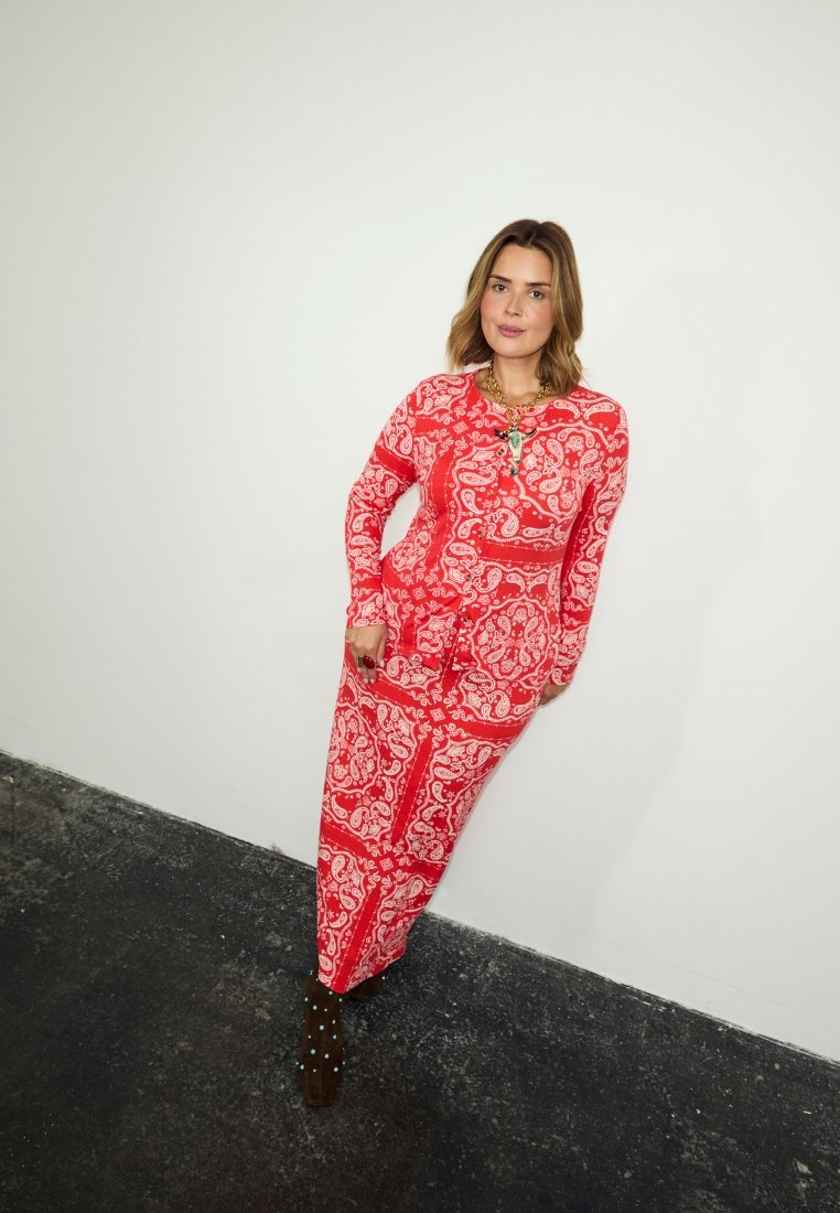 Γυναίκα που φοράει μακρύ φόρεμα κόκκινου με σχέδιο πεϊσλι, καφέ μποτάκια στον αστράγαλο με μπλε πουά και ένα χρυσό κολιέ, στέκεται μπροστά σε λευκό τοίχο.