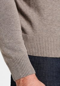 Beige stickad tröja med ribbade muddar och fåll, som visar ett strukturerat, mjukt tyg i närbild. I kombination med mörk denim.