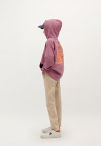 Paars hoodie met oranje grafisch ontwerp, beige broek en witte sneakers met een blauw logo en oranje accenten.