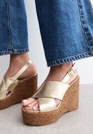 Gold wedge sandals with cork platform, crisscross metallic straps, open toe design, and adjustable ankle strap, set against denim fabric.