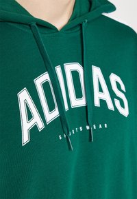 Grön hoodie i mjukt tyg med vit "ADIDAS"-logotyp och texten "SPORTSWEAR". Har en huva med dragsko och ribbad nederkant.
