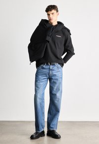 Svart genser med logo, brukt over skuldrene; blå jeans med avslappet passform; svarte lær sko; nøytral bakgrunn.