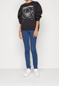 Černá mikina s bílým a tyrkysovým grafickým designem, v kombinaci s modrými úzkými džíny a bílými teniskami. Má volný střih a kulatý výstřih.