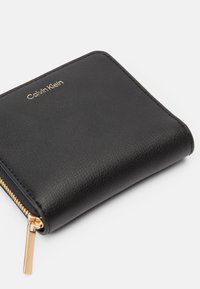 Fekete bőr Calvin Klein pénztárca, arany cipzárral és logóval a bal felső részen, zárt állapotban, fehér háttér előtt.