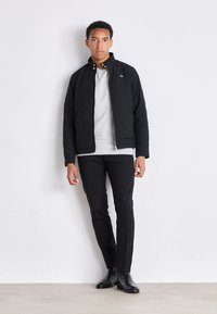 Zwarte gewatteerde jas met een staande kraag, ritsluiting, gedragen over een grijze sweatshirt, gecombineerd met zwarte slim-fit broek en zwarte leren laarzen.