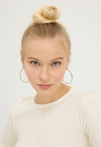 Blond haar in een knot, met grote zilveren oorringen, een geribbeld lichtbeige top met een ronde hals, tegen een witte achtergrond.